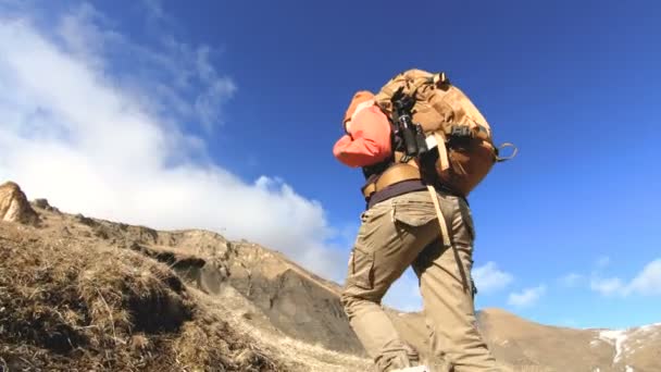 Gelukkige reiziger meisje fotograaf in zonnebril met een camera en een rugzak gaat bergopwaarts op een achtergrond van besneeuwde toppen op een zonnige dag — Stockvideo