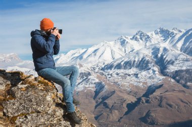 Güneş gözlüğü ve bir kap sakallı bir gezgin fotoğrafçı portresi dağların arka planında elinde ayna kamera ile bir kaya üzerinde oturur