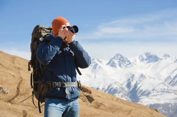Portrét vousatého mužského fotografa v brýlích a teplé sako s batohu na zádech a reflexní kamera v dlaních fotografuje na pozadí zasněžené hory — Stock fotografie