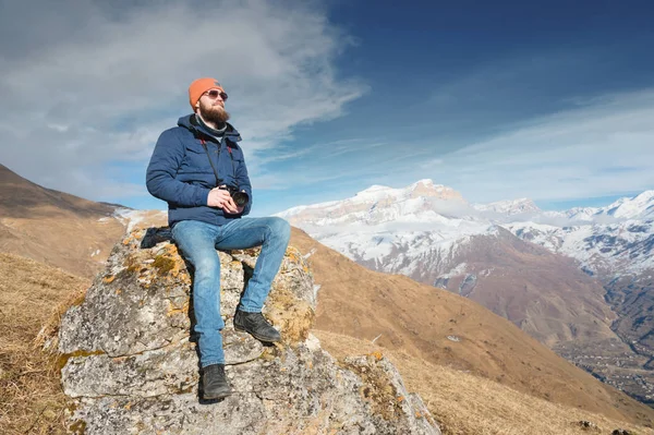 Portrait of a bearded traveler photographer in sunglasses and a cap sits on a rock with mirror camera in his hands against the backdrop of mountains — Stock Photo, Image