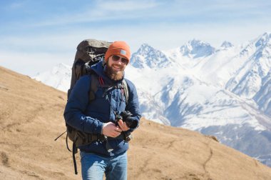 Güneşli bir günde karla kaplı dağların arka planına karşı güneş gözlüğü ile gülümseyen sakallı erkek fotoğrafçı portresi