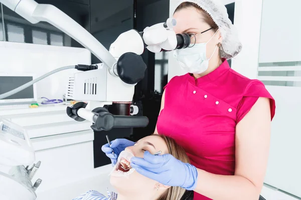 Μια γυναίκα επαγγελματίας οδοντίατρος εξετάζει μια γυναίκα ασθενή με ένα σταδολογικό μικροσκόπιο στο γραφείο της. Έννοια του επαγγέλματος του σταταολόγου — Φωτογραφία Αρχείου