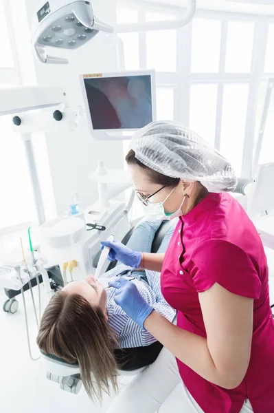 A professional dentist woman in glasses and overalls examines the oral cavity of a young girl in the dental chair using an intraoral stamotological video camera with LED illumination