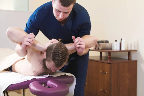 Un fisioterapeuta masculino hace un estiramiento de poder para un paciente masculino a un joven hipster barbudo en una sala de masajes médicos. Estirar la espalda y la columna vertebral —  Fotos de Stock