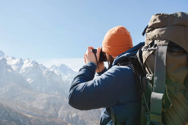 Portrét vousatého mužského fotografa v brýlích a teplé sako s batohu na zádech a reflexní kamera v dlaních fotografuje na pozadí zasněžené hory — Stock fotografie