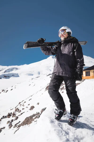 Retrato de Crescimento Esquiador macho barbudo envelhecido contra o fundo das montanhas do Cáucaso cobertas de neve. Um homem adulto usando óculos de esqui máscara e esquis capacete em seu ombro parece montanhas. Conceito de resort de esqui — Fotografia de Stock