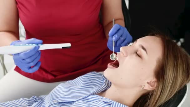 Uma dentista profissional de óculos e macacão examina a cavidade oral de uma jovem na cadeira dental usando uma câmera de vídeo estomatológica intraoral com iluminação LED — Vídeo de Stock