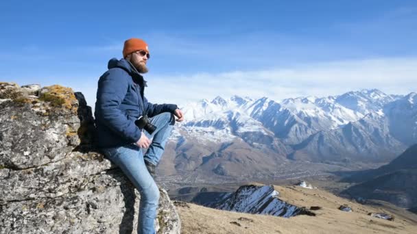 Porträt eines bärtigen Reisefotografen mit Sonnenbrille und Mütze sitzt auf einem Felsen mit Spiegelkamera in der Hand vor der Kulisse der Berge — Stockvideo