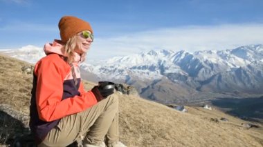 Elinde bir kupa ile şapka ve güneş gözlüğü güzel bir kız Portresi dağlarda bir taş üzerinde otururken kahve veya çay içiyor. Dağlarda turizm ve rekreasyon kavramı