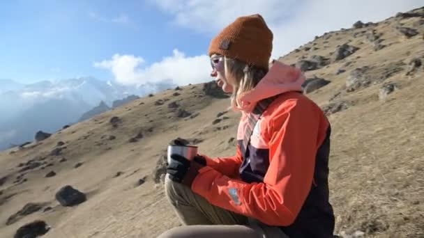 Porträtt av en vacker flicka i en hatt och solglasögon med en mugg i händerna dricker kaffe eller te medan du sitter på en sten i bergen. Begreppet turism och rekreation i fjällen — Stockvideo