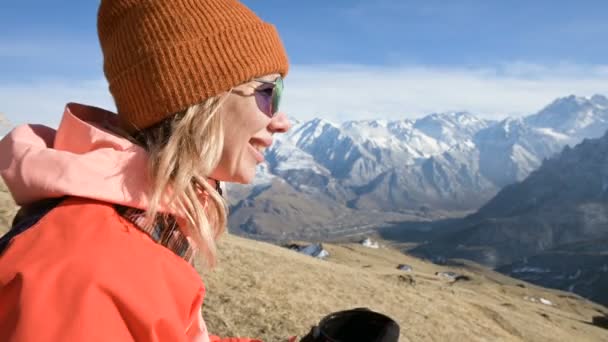 Retrato de uma menina bonita em um chapéu e óculos de sol com uma caneca em suas mãos está bebendo café ou chá enquanto sentado em uma pedra nas montanhas. O conceito de turismo e recreação nas montanhas — Vídeo de Stock