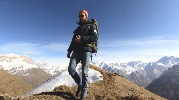 Счастливый путешественник бородатый мужчина фотограф в солнечных очках с камерой и рюкзаком идет в гору на фоне снежных пиков в солнечный день — стоковое видео