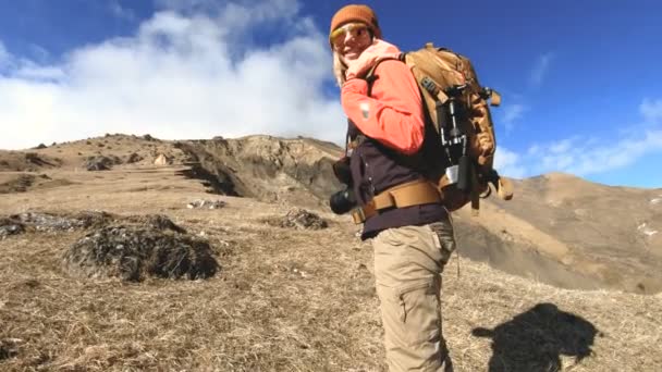 Feliz fotógrafa viajera en gafas de sol con una cámara y una mochila va cuesta arriba sobre un fondo de picos nevados en un día soleado — Vídeos de Stock