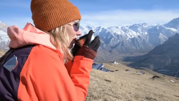 Retrato de uma menina bonita em um chapéu e óculos de sol com uma caneca em suas mãos está bebendo café ou chá enquanto sentado em uma pedra nas montanhas. O conceito de turismo e recreação nas montanhas — Vídeo de Stock