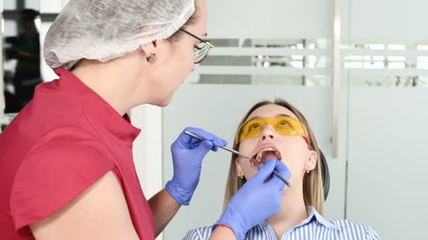 Koruyucu sarı gözlüklü güzel sarışın kız stamotolojist açık ağzını inceledi. Kadın diş hekimi bir diş hekimi aleti yardımıyla genç bir hastanın ağız boşluğu nu inceler — Stok video