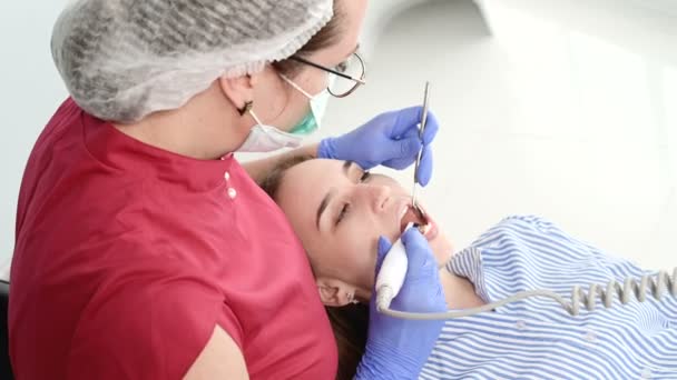Una dentista profesional en gafas y overoles examina la cavidad oral de una joven en la silla dental usando una cámara de video estamotológica intraoral con iluminación led — Vídeo de stock