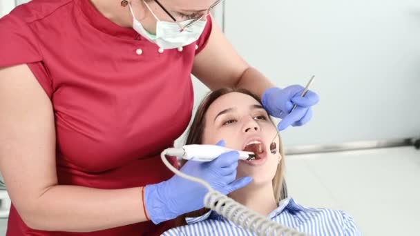 Профессиональная стоматологическая женщина в очках и комбинезонах осматривает полость рта молодой девушки в стоматологическом кресле с помощью внутриротовой стоматологической видеокамеры со светодиодной подсветкой — стоковое видео