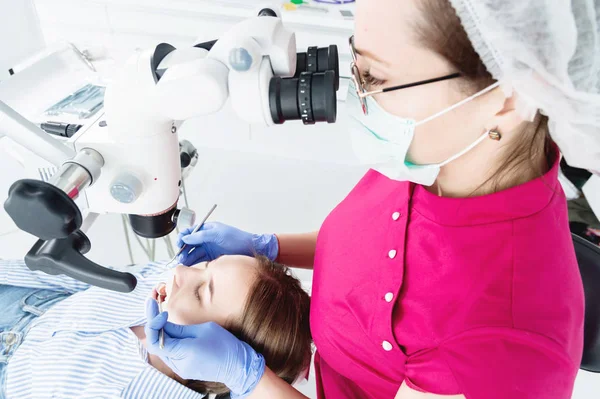 Μια γυναίκα επαγγελματίας οδοντίατρος εξετάζει μια γυναίκα ασθενή με ένα σταδολογικό μικροσκόπιο στο γραφείο της. Έννοια του επαγγέλματος του σταταολόγου — Φωτογραφία Αρχείου