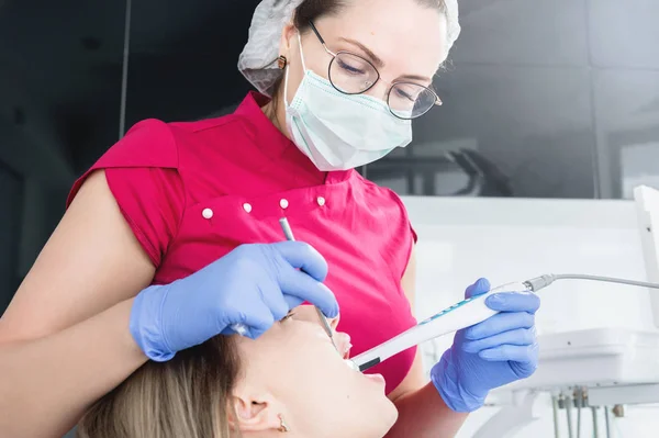 Uma dentista profissional de óculos e macacão examina a cavidade oral de uma jovem na cadeira dental usando uma câmera de vídeo estomatológica intraoral com iluminação LED — Fotografia de Stock