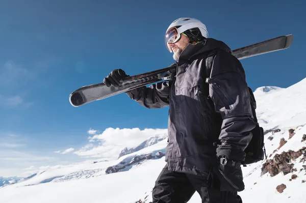 Close-up Ritratto barbuto sciatore di sesso maschile invecchiato sullo sfondo delle montagne. Un uomo adulto che indossa maschera da sci googles e sci casco sulla spalla guarda montagne. Concetto di stazione sciistica — Foto Stock