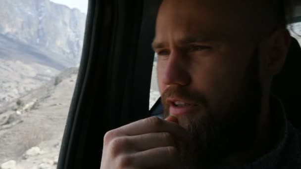 Portrait en gros plan d'un hipster barbu parlant assis dans la voiture sur le siège arrière et méditant sur les côtés sur fond de montagnes — Video