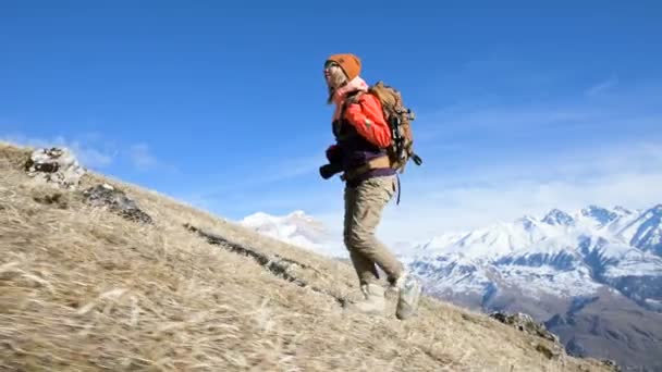 Hermosa chica fotógrafa en un sombrero y gafas de sol con una gran mochila y una cámara alrededor de su cuello sube la colina sobre el fondo de las montañas cubiertas de nieve — Vídeos de Stock