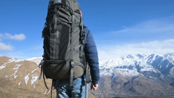 Um homem barbudo de chapéu de óculos de sol com uma mochila e uma câmera fica no alto das montanhas e olha de lado contra o pano de fundo das montanhas cobertas de neve — Vídeo de Stock