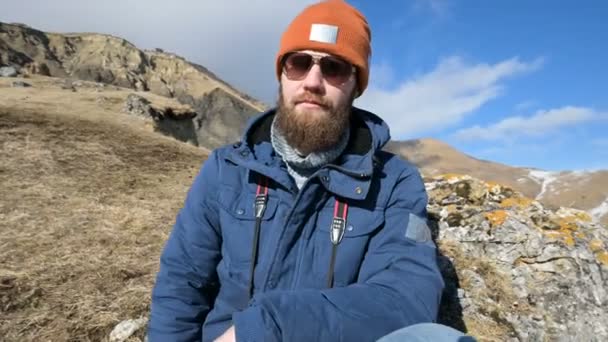 Portré egy szakállas utazó fotós napszemüveg és egy sapka ül egy szikla tükörkamerával a kezében a háttérben a hegyek — Stock videók
