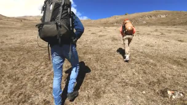 Zwei Touristenfotografen mit Rucksäcken in Hüten und Sonnenbrillen steigen mit Kameras in der Hand vor dem Hintergrund verschneiter Berge auf den Hügel im gelben Gras. Zeitlupe — Stockvideo