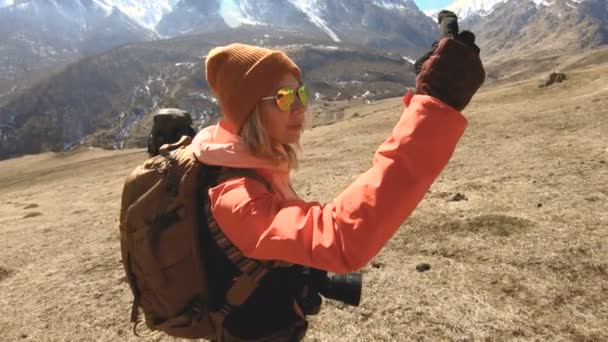 Портрет щасливої дівчини мандрівника-фотографа в капелюсі і сонцезахисних окулярах з камерою навколо шиї і телефоном в руках на тлі гір фотографує на своєму смартфоні — стокове відео