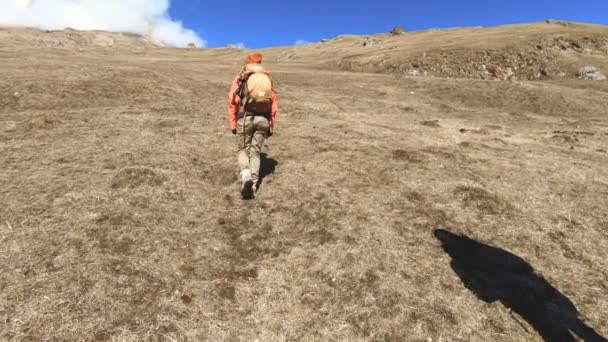 Dos fotógrafos turistas con mochilas en sombreros y gafas de sol suben la colina en la hierba amarilla con cámaras en sus manos contra el fondo de las montañas nevadas. cámara lenta — Vídeos de Stock