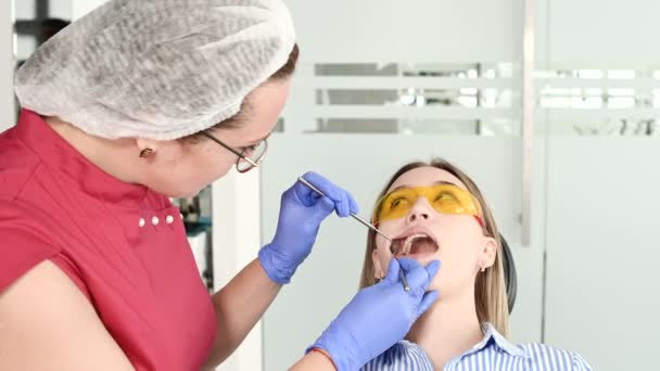 スタモトロジストの保護黄色のメガネをかけたかわいいブロンドの女の子は、彼女の開いた口を調べました。女性歯科医は、歯科医の器具の助けを借りて若い患者の口腔を検査する — ストック動画
