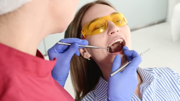 Menina loira bonita em óculos protetores amarelos no stamotologist examinou sua boca aberta. Dentista feminina examina a cavidade oral de um paciente jovem com a ajuda de um instrumento de dentista — Vídeo de Stock