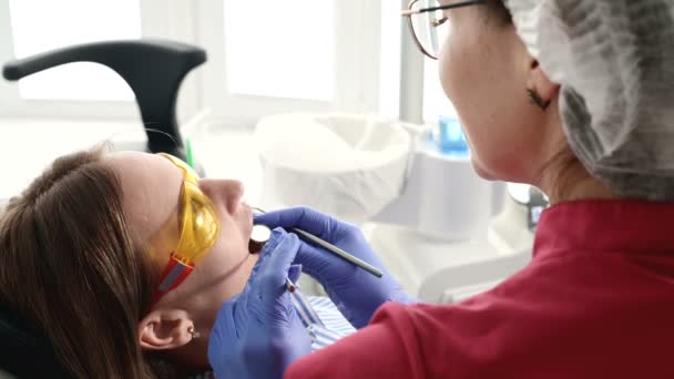 スタモトロジストの保護黄色のメガネをかけたかわいいブロンドの女の子は、彼女の開いた口を調べました。女性歯科医は、歯科医の器具の助けを借りて若い患者の口腔を検査する — ストック動画
