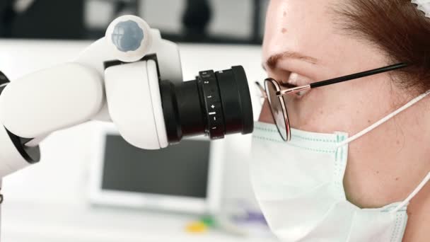 Κοντινό-up μια γυναίκα με γυαλιά με έναν επαγγελματία οδοντίατρο που συνεργάζεται με ένα επαγγελματικό στανολογικό μικροσκόπιο στο γραφείο της. Έννοια του επαγγέλματος του σταταολόγου — Αρχείο Βίντεο