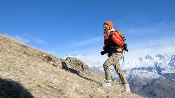Vacker flicka fotograf i en hatt och solglasögon med en stor ryggsäck och en kamera runt halsen klättrar upp för backen mot bakgrund av snötäckta berg — Stockvideo