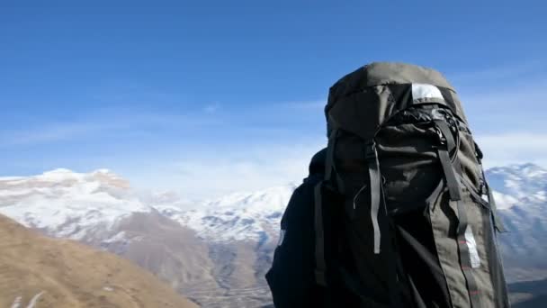 サングラスをかけたひげを生やした男がリュックサックとカメラを持って山の高く立ち、雪に覆われた山々を背景に横を見る — ストック動画