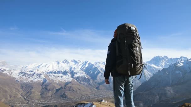 Un homme barbu en chapeau de lunettes de soleil avec un sac à dos et une caméra se tient haut dans les montagnes et regarde sur les lignes de touche dans le contexte des montagnes enneigées — Video