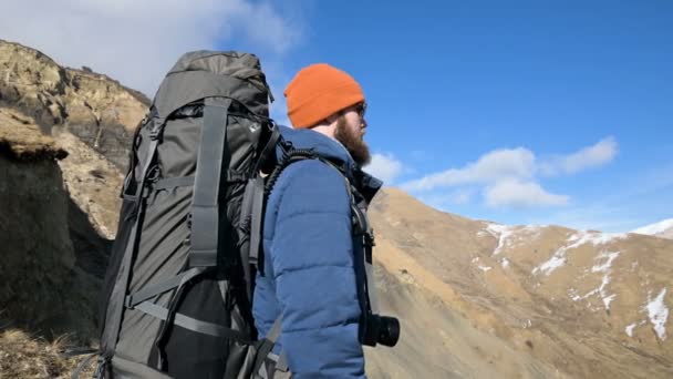 Бородатый мужчина в шляпе в солнечных очках с рюкзаком и камерой стоит высоко в горах и смотрит в сторону на фоне заснеженных гор — стоковое видео