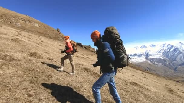 Due fotografi turisti con zaini in cappelli e occhiali da sole salgono sulla collina sull'erba gialla con le macchine fotografiche in mano sullo sfondo di montagne innevate. rallentatore — Video Stock