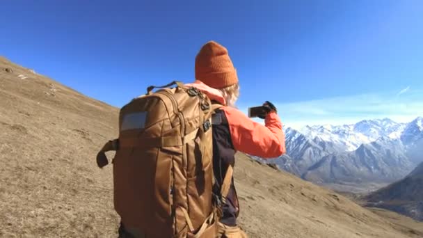 Retrato de una fotógrafa viajera feliz con sombrero y gafas de sol con una cámara alrededor del cuello y un teléfono en las manos en el fondo de las montañas toma fotos en su teléfono inteligente — Vídeo de stock