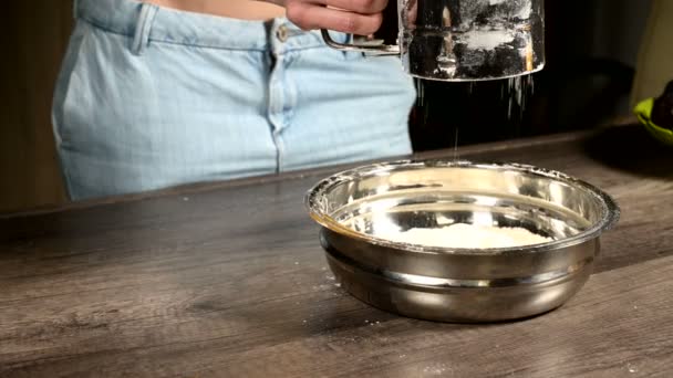 Κοντινό κομμάτι ενός γυναικείου χεριού που κοσκινίζει το αλεύρι με μια κούπα κόσκινο σε ένα μεταλλικό μπολ στην κουζίνα της οικίας. Μαγειρική σπίτι ψήσιμο — Αρχείο Βίντεο