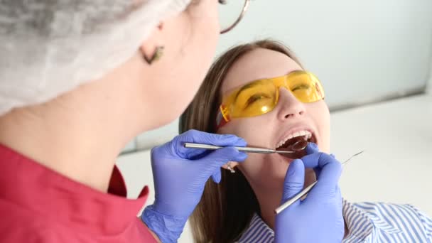 漂亮的金发女孩在保护黄色眼镜上的葡萄球菌学家检查她张开的嘴。女牙医在牙医仪器的帮助下检查年轻病人的口腔 — 图库视频影像