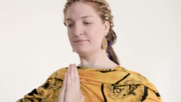 En flicka med dreadlocks och händer i armband i en gest av Namaste och Namaskar. Flickan i en autentisk halsduk öva meditation stående i studion på en vit bakgrund — Stockvideo