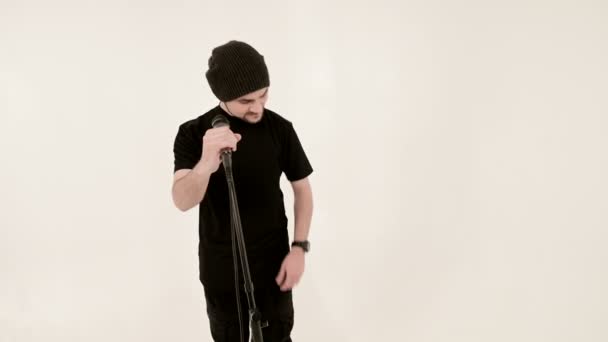 Frontman zanger rock pop met een stijlvolle baard in zwarte kleding en een hoed met een microfoon in zijn handen die expressief agressief zingen in de studio tegen de achtergrond van witte muren — Stockvideo