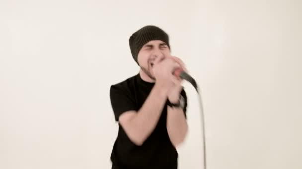 Vocalista pop rock Frontman com uma barba elegante em roupas pretas e um chapéu com um microfone em suas mãos cantando expressivamente agressivamente no estúdio contra o fundo das paredes brancas — Vídeo de Stock