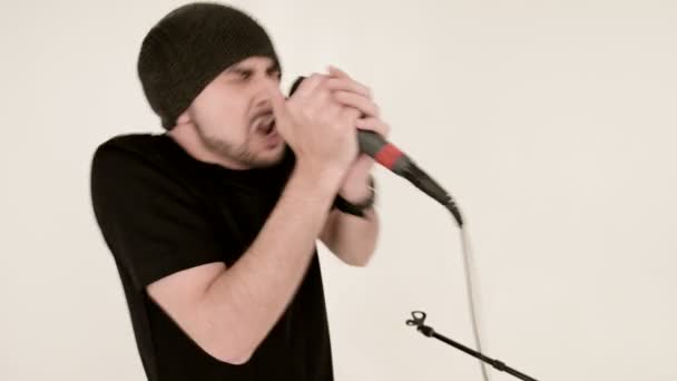 Frontman zanger rock pop met een stijlvolle baard in zwarte kleding en een hoed met een microfoon in zijn handen die expressief agressief zingen in de studio tegen de achtergrond van witte muren — Stockvideo