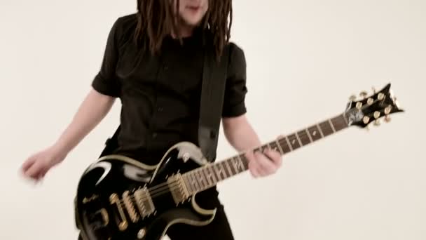 Elegante guitarrista solo com dreadlocks em sua cabeça e em roupas pretas em um fundo branco tocando expressivamente a guitarra preta em um estúdio branco — Vídeo de Stock
