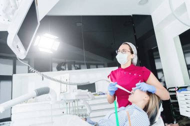 Gözlük ve tulum profesyonel bir diş hekimi kadın Led aydınlatma ile bir intraoral stamotolojik video kamera kullanarak diş sandalyesinde genç bir kızın ağız boşluğu inceler