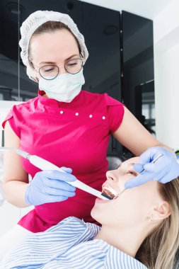 Gözlük ve tulum profesyonel bir diş hekimi kadın Led aydınlatma ile bir intraoral stamotolojik video kamera kullanarak diş sandalyesinde genç bir kızın ağız boşluğu inceler